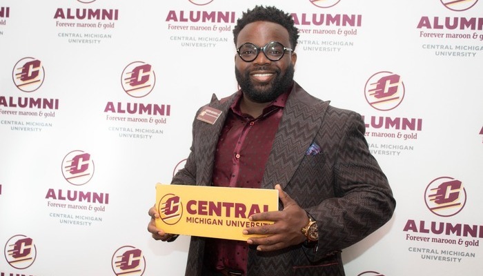 alumni_fuelyourpride_700x400 A CMU alum with glasses and a beard holds a gold sign reading 'Central Michigan University', against a backdrop featuring CMU Alumni branding.