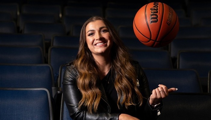 A woman in a black leather jacket smiles and tosses a basketball in the air while sitting.