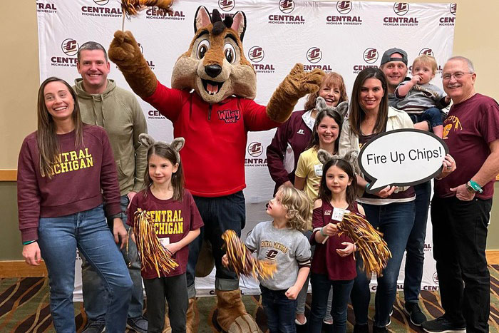 A multigenerational family, wearing CMU apparel, posing with a mascot in front of a CMU backdrop. One person holds a speech bubble that says 'Fire Up Chips!'