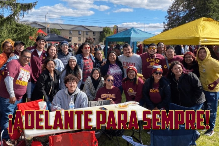 A large group of people stand outdoors at the 2022 Latine Alumni Tailgate. There is text on the image that state Adelante para sempre which translates to Fire up forever.
