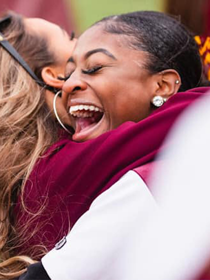 This is a photo of two women embracing in a hug. One wearing maroon with long blonde hair and one wearing white with black hair.