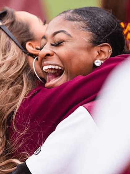 This is a photo of two women embracing in a hug. One wearing maroon with long blonde hair and one wearing white with black hair.