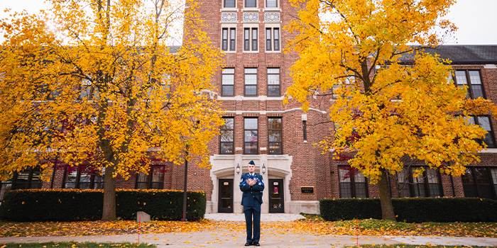 ROTC alumni chapter main page photo 700x350 A person in a military uniform stands on a sidewalk in front of a building.