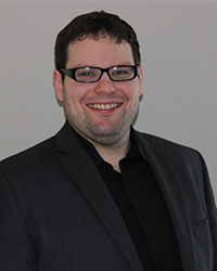Jason Dizik's previous headshot for the Alumni Awards where he is wearing a black button down shirt and black blazer and has short brown hair and black glasses while smiling with teeth.