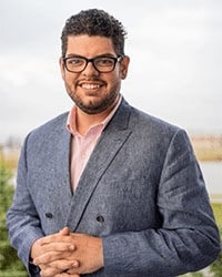 Zach Huffman's headshot for the Alumni Awards where he is wearing a gray suit jacket with a pink button-down shirt underneath and black glasses with short brown hair and he is smiling with teeth.