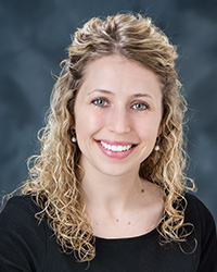 A woman with blonde shoulder length curly hair, half hair is pulled back, green/blue eyes, light pink lipstick, smile with teeth, dangling pearl earrings, swoop neck black shirt, and gray background.