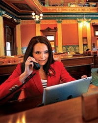 Andrea LaFontaine's headshot for the Alumni Awards where she is on the phone while staring at a computer as she is wearing a bright red blazer and has long dark brown hair in a government building.
