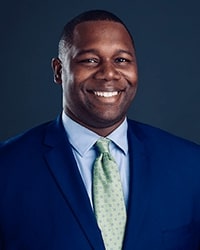 A man with short black hair, smile with teeth, dark brown eyes, light blue button up dress shirt with a collar, light green long tie with spots, navy blue suit coat, and gray background.