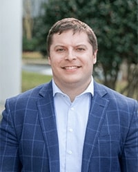 Steve McIsaac's previous headshot for the Alumni Awards where he is wearing a light blue button-down shirt with a dark blue suit jacket that has light blue stripes and he has short brown hair and is smiling with teeth.