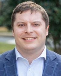 Steve McIssac wearing a light blue colored button down shirt with a color, dark blue dress coat with light blue stripes, short brown hair, outdoor greenery background.