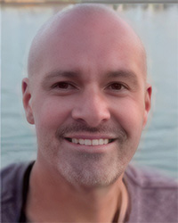 A man with a gray beard, smile teeth, maroon short sleeve shirt, dark brown eyes, light blue water background.