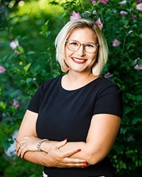 This is a headshot photo of Stephanie wearing a black top in front of greenery outdoors.