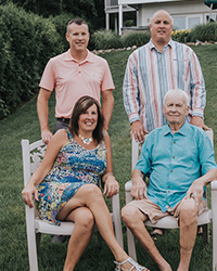 This is a family photo of four individuals. Two are seated in chairs in the first row; a woman to the left and a man to the right. In the back row, there are two standing men.