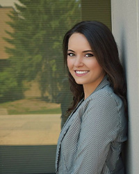 Headshot of 2024 Alumni Award 10 Within 10 Recipient Sabrina Bierstetel standing to the side wearing a blazer with thin black and white stripes and she has long brown hair and is smiling with teeth.