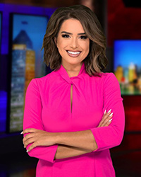 Headshot of 2024 Alumni Awards 10 Within 10 recipient Erica Francis and she is wearing a bright pink dress and has shoulder-length brown and is standing and smiling in front of a news station background.