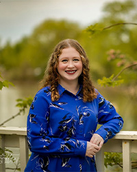 Headshot of 2024 Alumni Awards 10 Within 10 Recipient Leah Maskovitz where she is standing against a wooden railing wearing a blue long-sleeve dress and she has shoulder-length curly hair and is smiling with teeth.