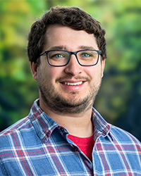 Headshot of Matthew Tuftedal for the 2024 10 Within 10 Alumni Awards and he is wearing a blue and red plaid shirt, has short brown hair and glasses, and is smiling with teeth.