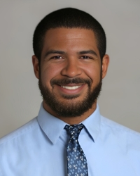 A man with short dark hair and a full beard is smiling at the camera. He is wearing a light blue dress shirt and a blue patterned tie. The background is a plain, neutral color.