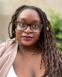 A woman with tortoiseshell glasses looks at the camera. She is wearing a light-colored top, a small gold necklace, and dark lipstick. Green foliage is blurred in the background.
