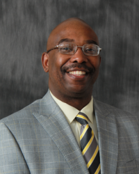 Damon Brown 200x250 A man with a bald head wears eyeglasses, a grey suit and striped tie.