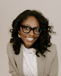 Dyese Matthews 200x250 A woman with dark, curly hair wears glasses and a tan blazer over a white shirt.