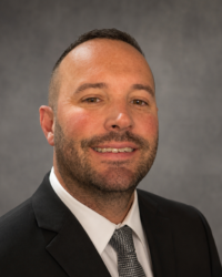 John Hunter 200x250 Headshot of a man with a short beard and short dark hair is wearing a black suit, dark tie and white dress shirt.