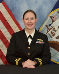 Lydia Sankey 200x250 A woman in a military uniform smiles with one hand on top of the other in front of her. A US flag is over her left shoulder and a white flag with various brown symbols including a small US flag on it is over her right shoulder.