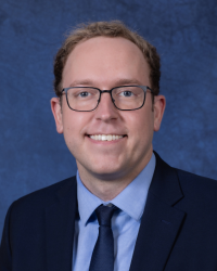 Matthew Boak 200x250 A man with glasses and short brown hair smiles while wearing a dark suit, navy blue tie and light blue shirt.