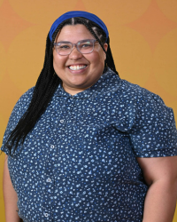 Sam Jenkins 200x250 A woman wearing glasses with long black hair, a royal blue headband and a blue shirt with light blue spots all over it is smiling at the camera.