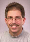 Haxer This is a headshot of a man wearing a gray mock neck shirt with light brown hair a mustache and glasses.