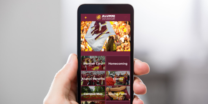 alumni_connectcmu_700x350 A hand holds a smartphone displaying the homescreen of the CMU Alumni App, featuring a banner image and menu options like Member Card, Homecoming, Alumni Benefits, Events, Campus Map and Giving Form.