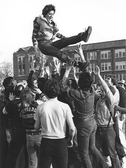 A group of people are gathered close together and toss a person in the air with arms outstretched to catch them.