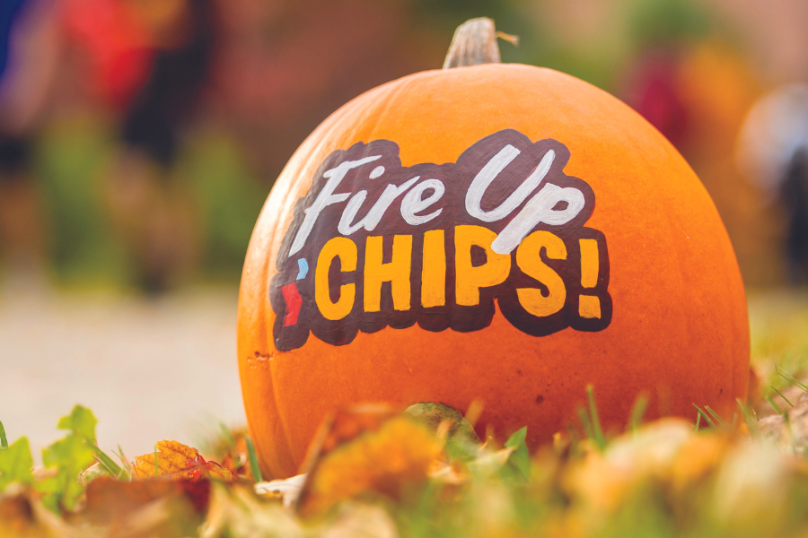 A pumpkin on the ground outdoors that is painted with the words Fire Up Chips.