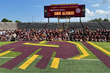 Over 300 Central Michigan University alumni posing together during alumni takeover weekend on the football field in front of the scoreboard.