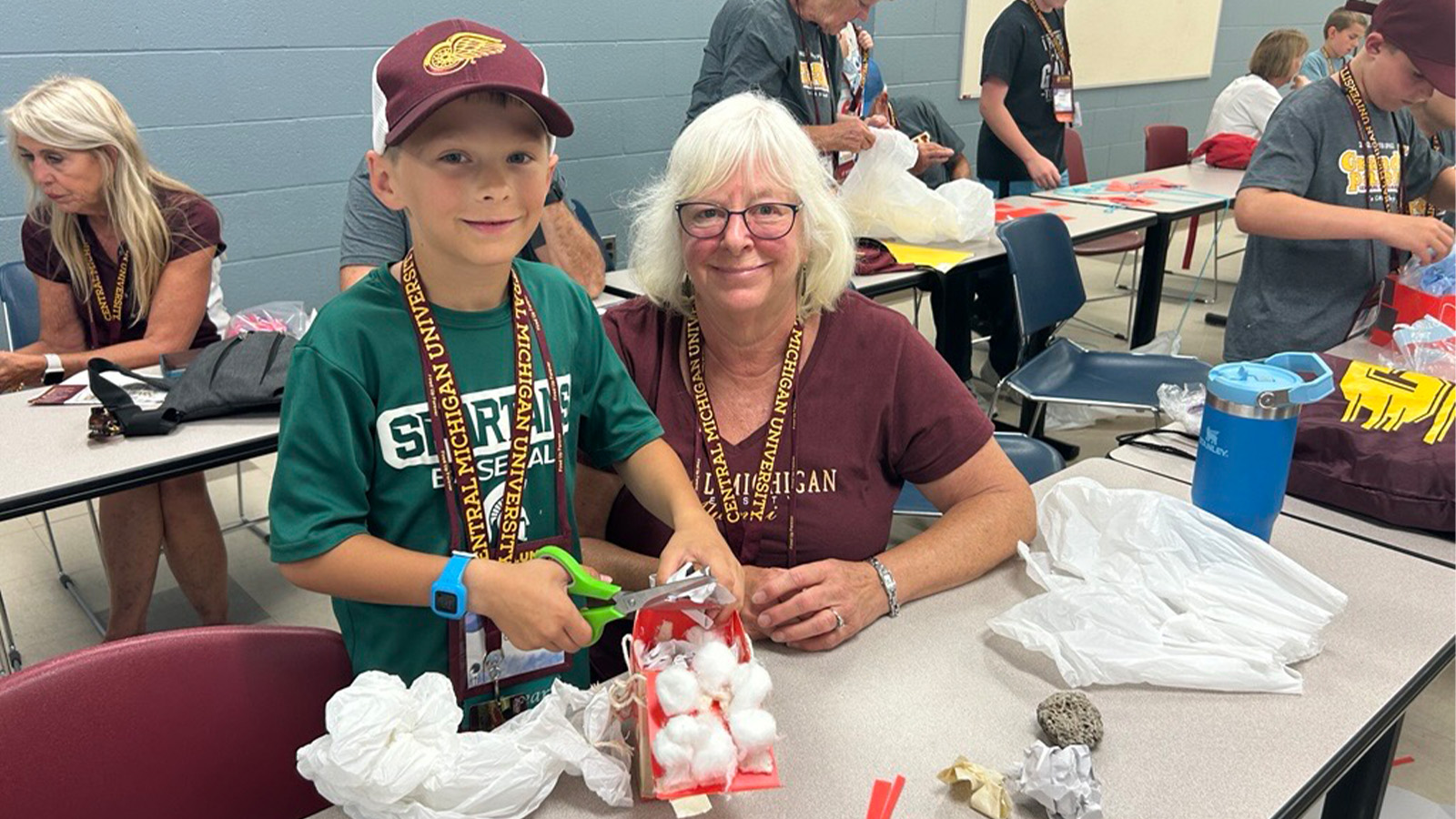 A woman and a boy smile while working on a craft project in a classroom. The boy has scissors in his hands.
