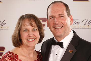 Woman in red dress with short brown hair smiles while posing with a man who has short dark hair and is wearing a tuxedo.