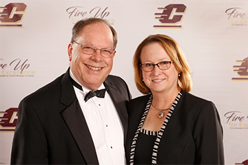 A man in a tuxedo smiles and poses next to a smiling woman wearing a black dress.
