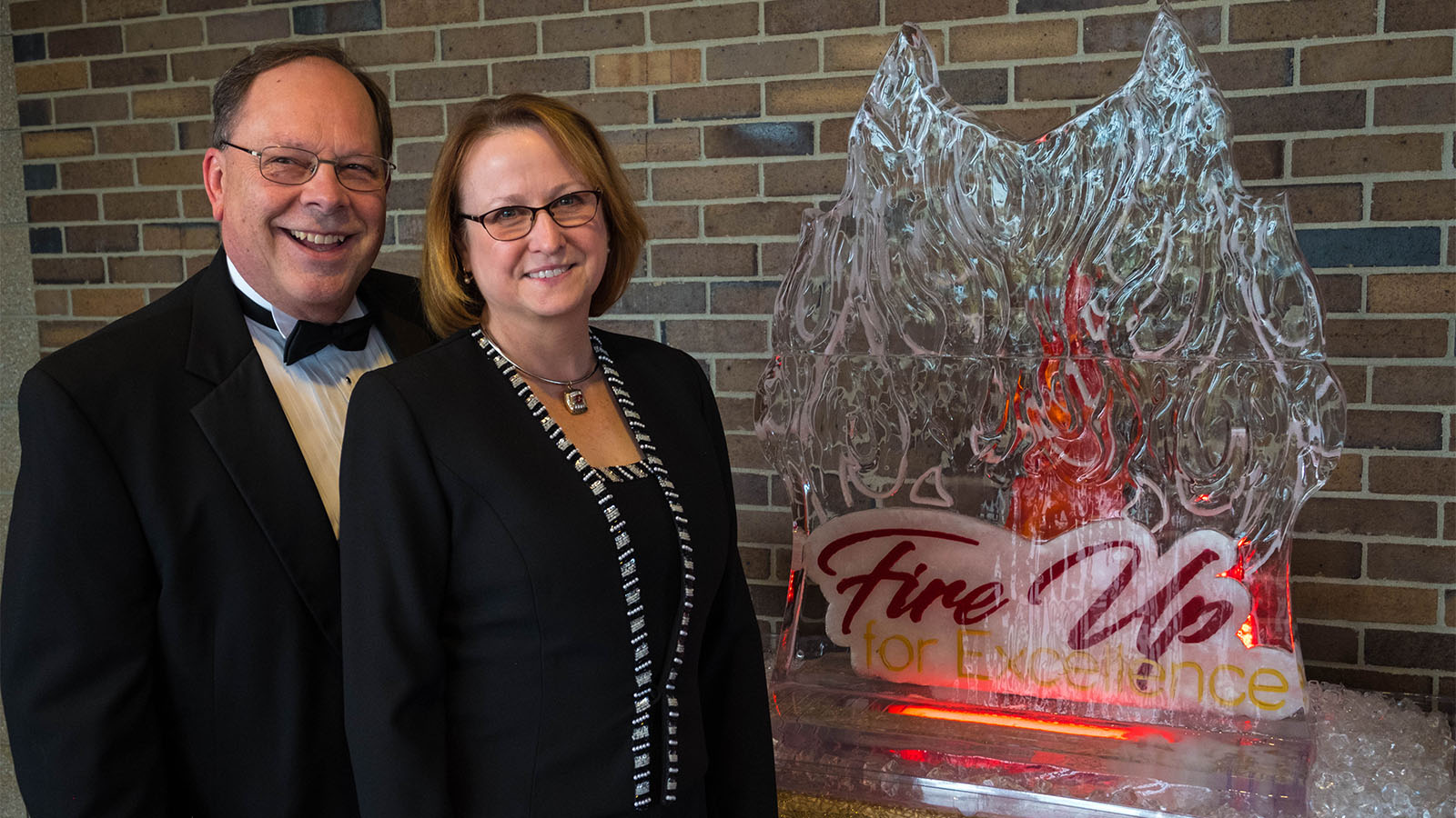 A man and woman standing next to a carved ice block with a logo inside that reads Fire Up for Excellence.