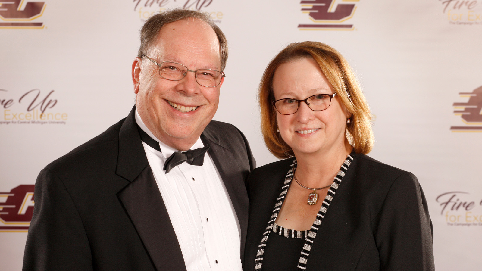 A man in a tuxedo smiles and poses next to a smiling woman wearing a black dress.