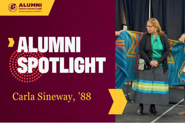 A Central Michigan University Alumni Spotlight graphic featuring Carla Sineway, '88. Carla is wearing a green top and long patterned skirt, standing in front of a blue and gold star quilt being held up behind her.