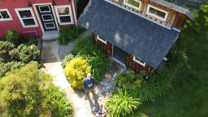 Aerial view of John Nelson, holding a mug outside his custom pine backyard office shed surrounded by landscaping.