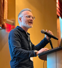 A silver-haired man with glasses wearing a blue long-sleeve shirt speaking at a podium with a microphone.