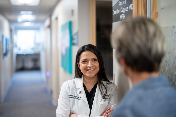 CMU alumnus Dr. Kristen Carrillo-Kappus, wearing an OB/GYN white lab coat while speaking with a colleague in a medical clinic hallway.