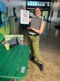A smiling woman with red hair wearing cargo pants next to a green picnic table, holding up a flyer and a gray presentation board.