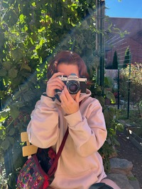 A woman wearing a light pink hoodie holding a vintage camera up to their face to take a photo, with green foliage in the background.