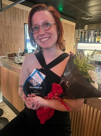 A smiling woman with red hair wearing glasses and a black dress, holding an award plaque and a bouquet of flowers indoors.
