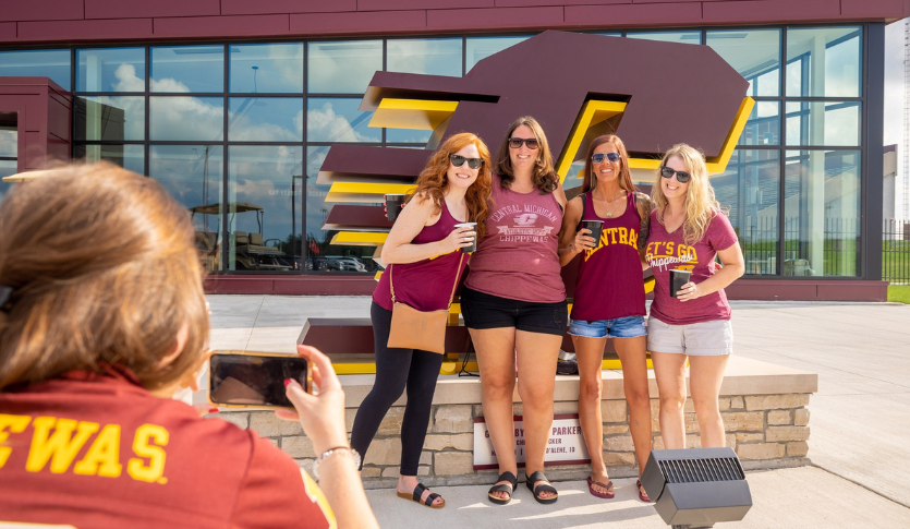 This is an image of one alumna taking a photo of four alumnae, all wearing maroon tops, standing in front of the Action C statue located in front of the Chippewa Champion Center.