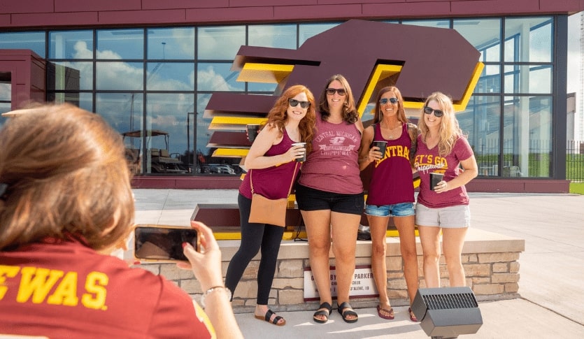 This is an image of one alumna taking a photo of four alumnae, all wearing maroon tops, standing in front of the Action C statue located in front of the Chippewa Champion Center.