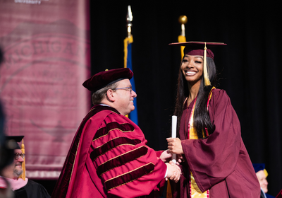 A woman crosses the stage in her cap and gown and receives her diploma from President Davies.