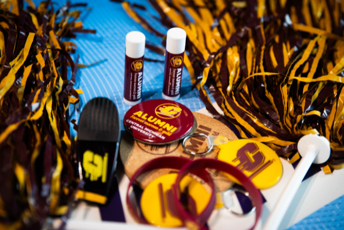 CMU Alumni swag scattered on a table, including maroon and gold pom poms, buttons, and keychains.